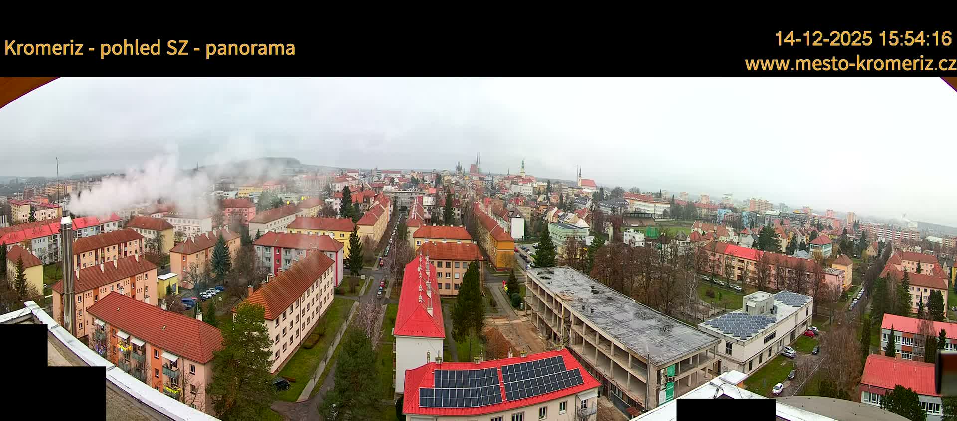 A wide, panoramic view of a sprawling city on an overcast day, featuring a dense mix of residential buildings, prominent church spires in the distance, a large partially constructed building in the foreground, and multiple plumes of smoke rising into the grey sky, with bare trees suggesting a cooler season.