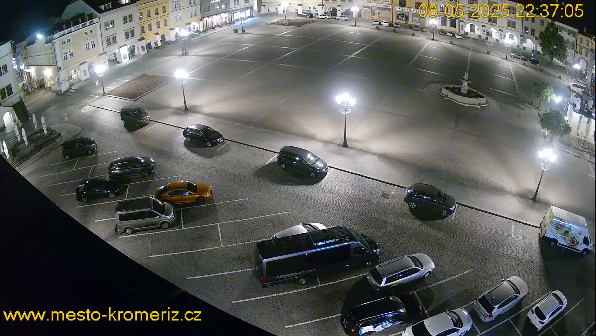 A nighttime aerial view of a town square with several cars parked in a lot surrounding a central fountain, under clear skies.