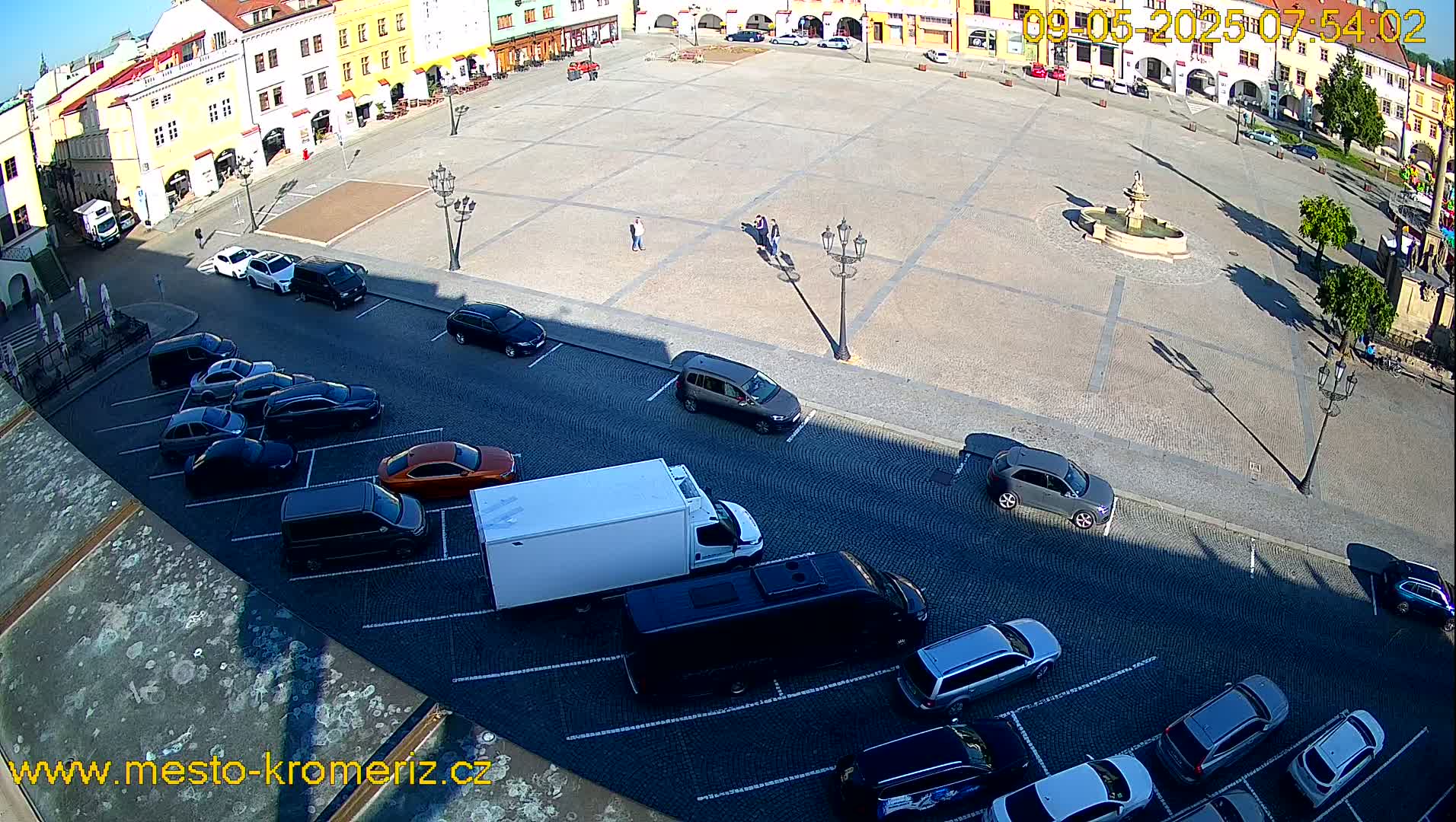 A sunny day reveals a town square with a fountain, surrounded by buildings and a parking lot filled with cars and a van.