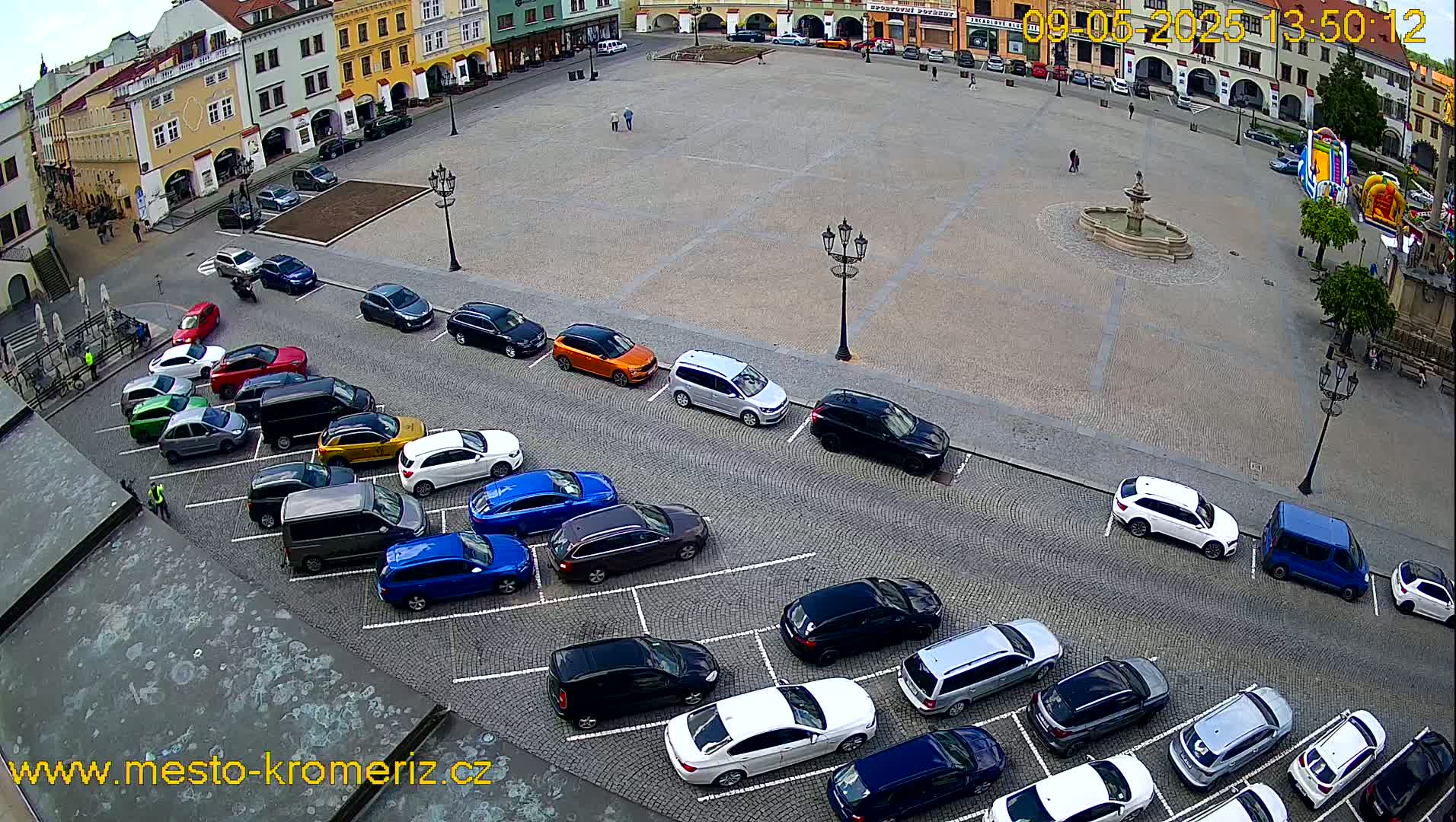 An aerial view shows a town square with a fountain and several cars parked along the sides under a partly cloudy sky.