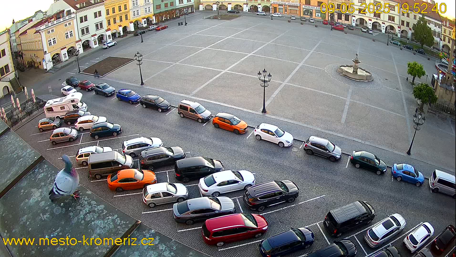 A large town square with a fountain in the center is surrounded by buildings and cars parked along its perimeter under a partly sunny sky.