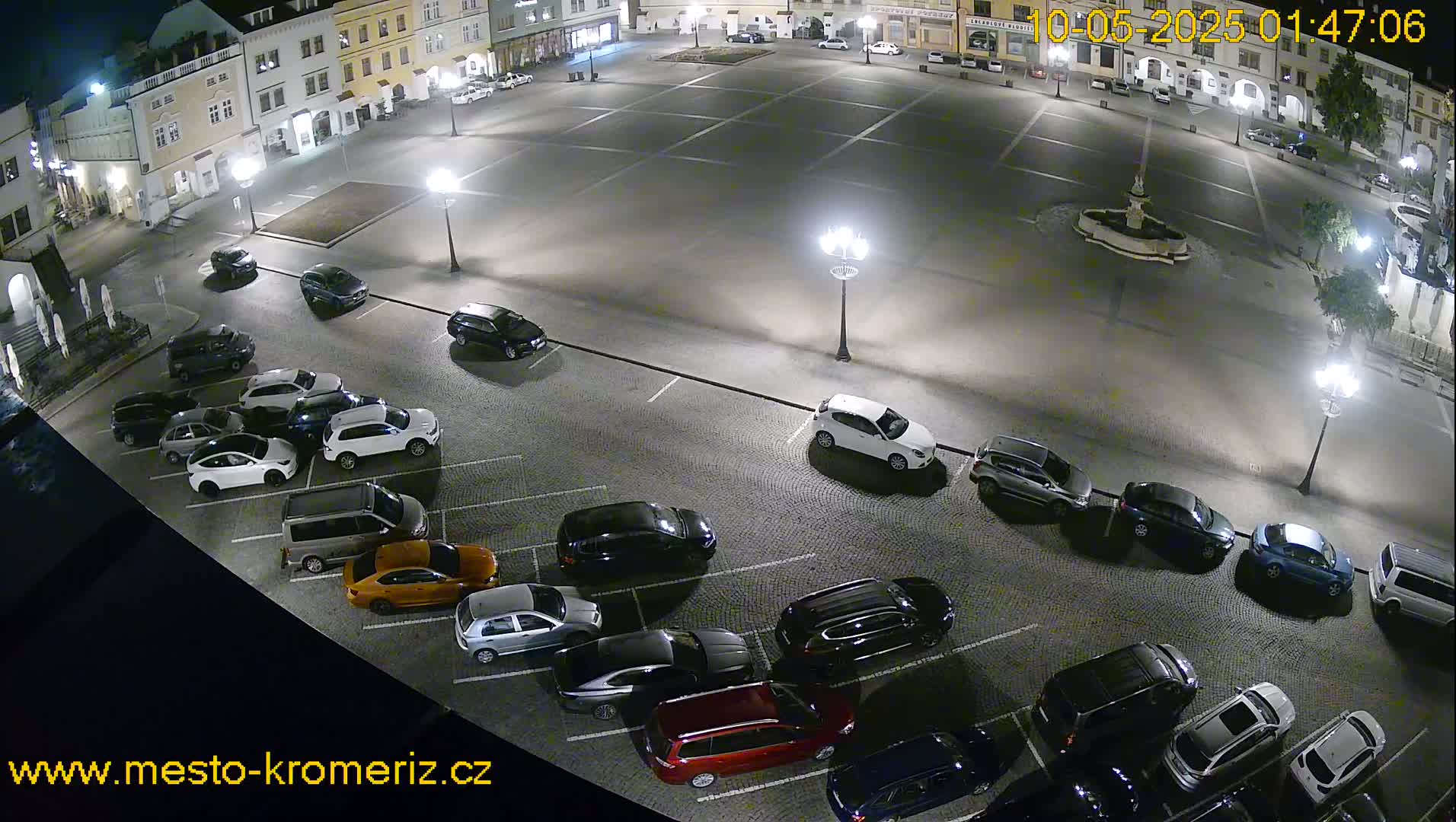 A nighttime aerial view of a town square shows numerous cars parked in a lot surrounding a fountain, under bright streetlights.