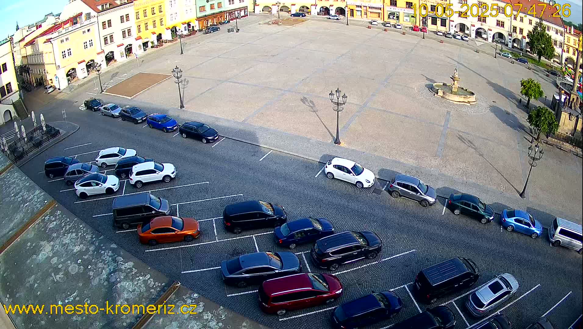 A large town square, mostly empty except for cars parked along the sides, features a central fountain and is bathed in sunlight.