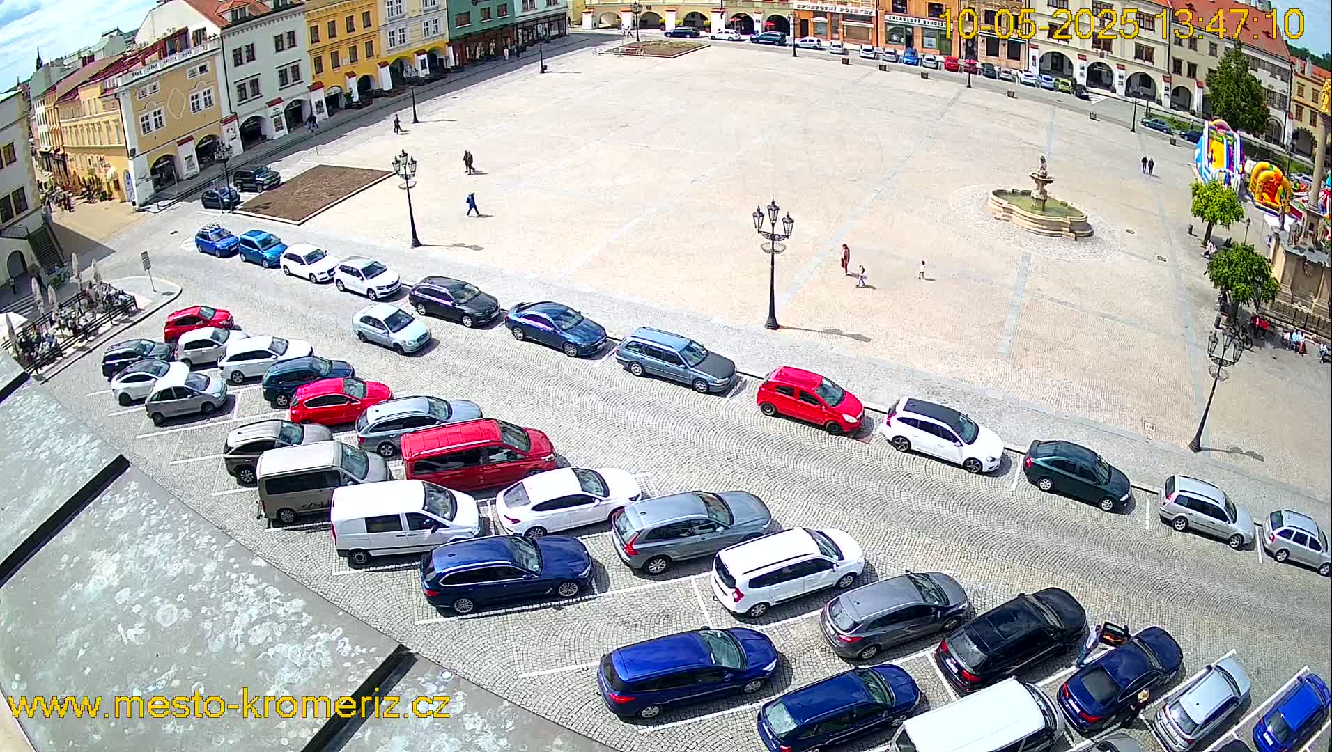 A sunny day shows a town square with a fountain and several cars parked along the side of the square.