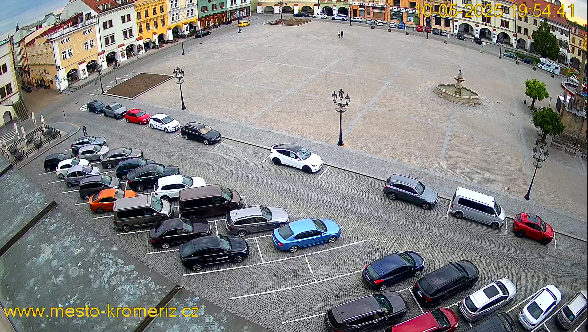 A large town square with a fountain in the center is surrounded by buildings and lined with parked cars.