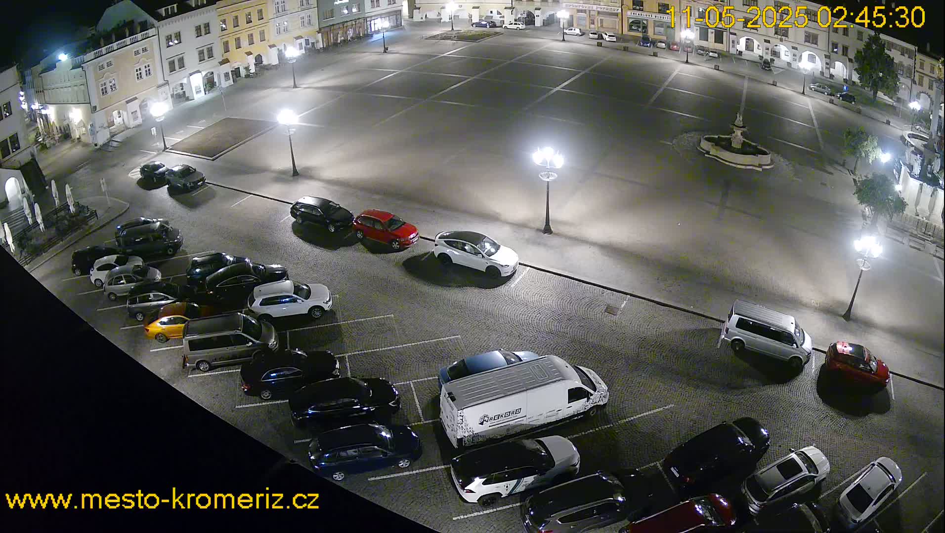A nighttime aerial view of a town square shows a mostly empty plaza with cars parked along the edges, illuminated by streetlights.