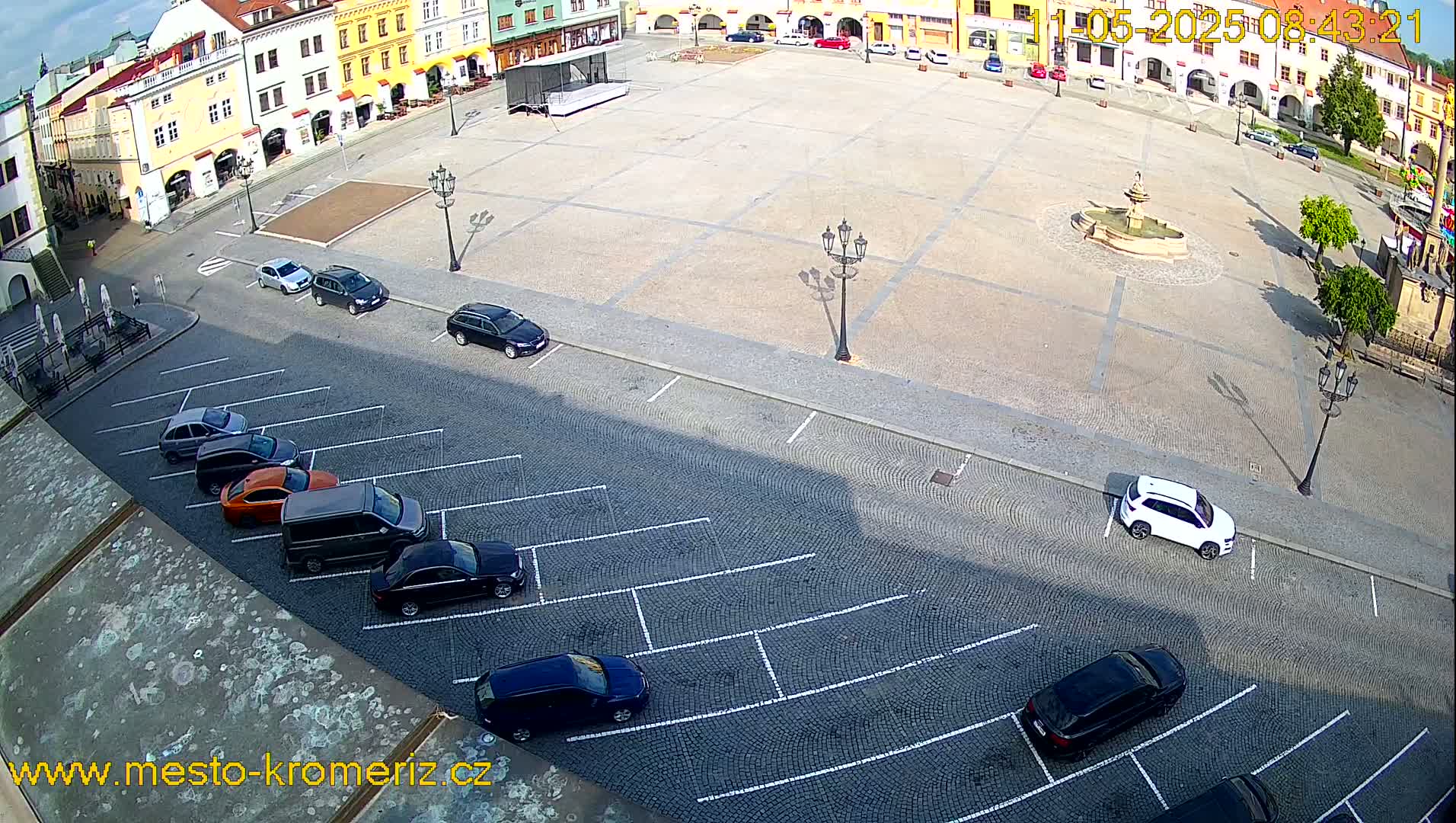 A mostly empty cobblestone town square with a fountain at its center and several cars parked along the perimeter on a sunny day.