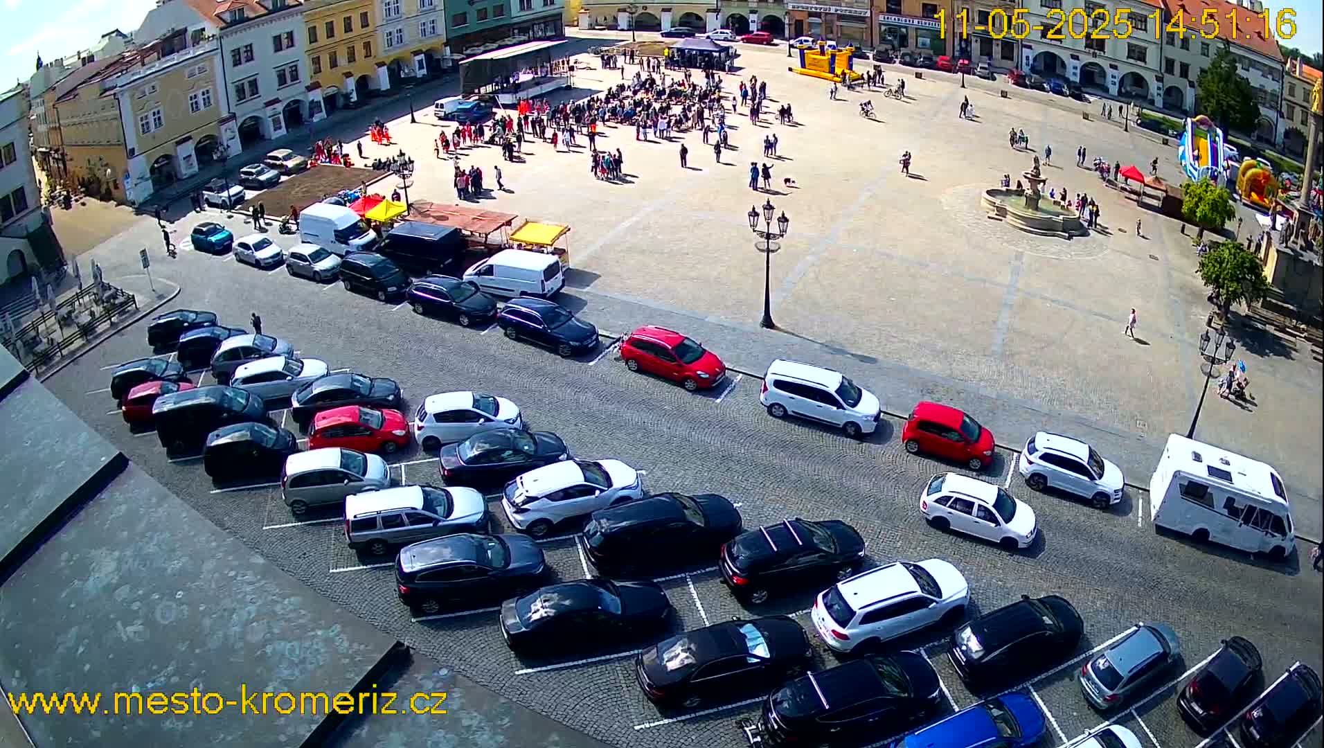 A town square with many people gathered, a fountain, and several cars parked in a lot nearby under a sunny sky.