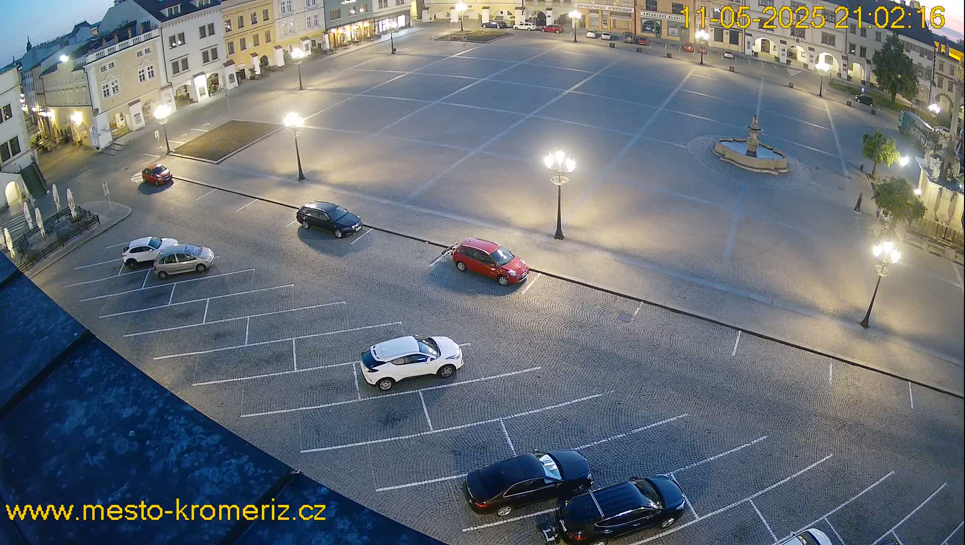 A nighttime, high-angle view of a town square's parking lot with several cars parked and a large, empty square illuminated by streetlights.