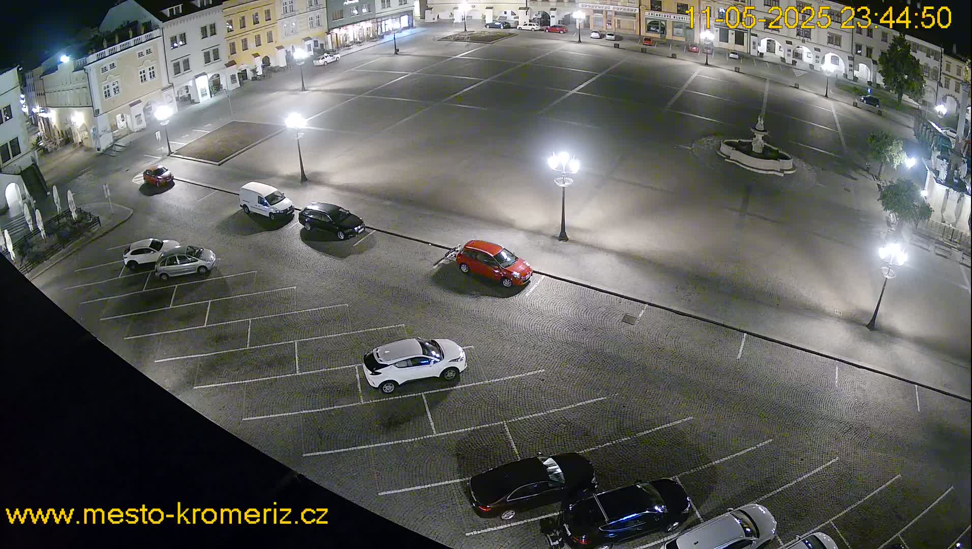 A nighttime, overhead view of a town square's parking lot with several cars parked, and a fountain in the center of the square.
