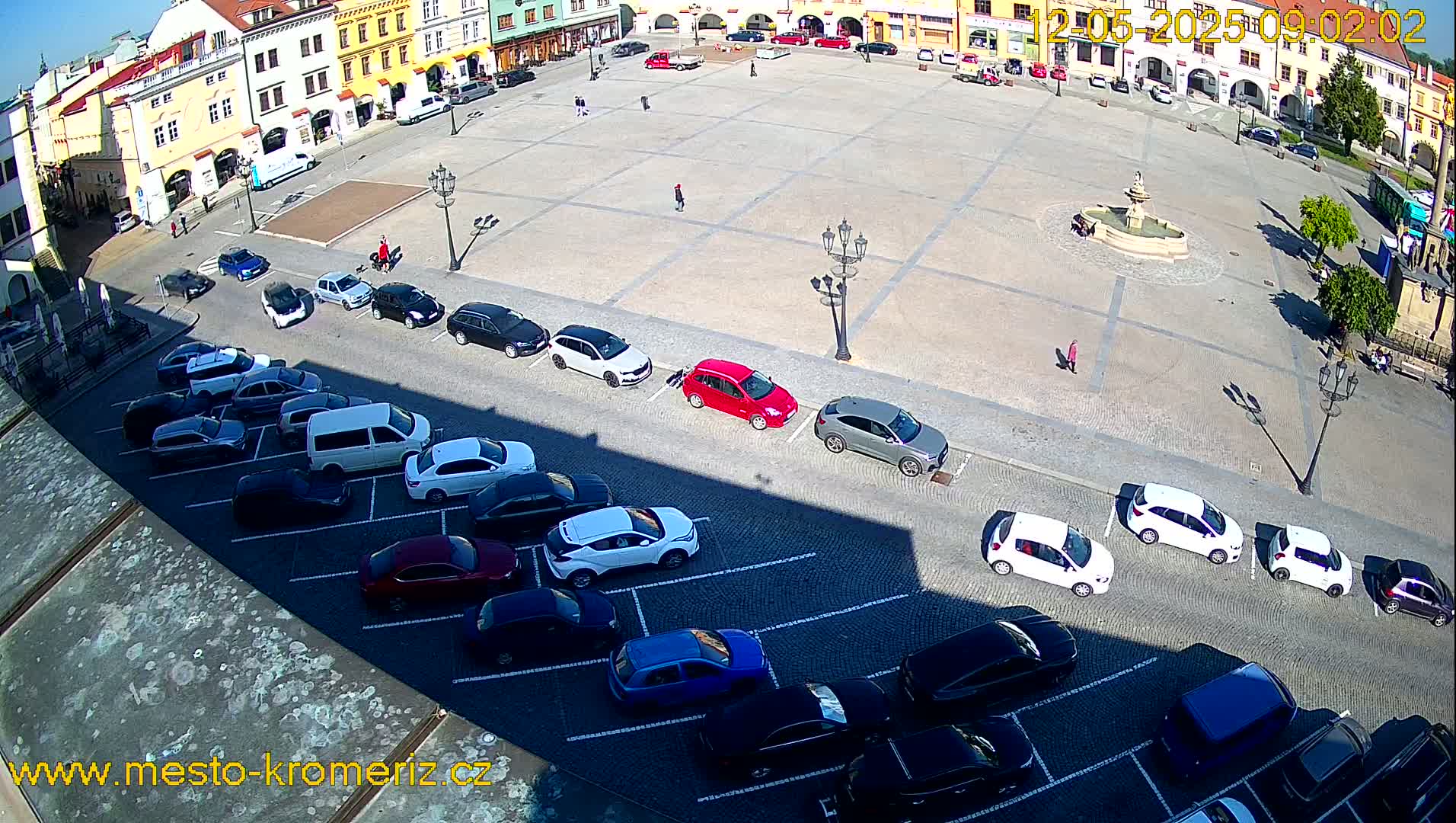 A sunny day reveals a large town square with cars parked along one side and a fountain in the center, surrounded by historical buildings.