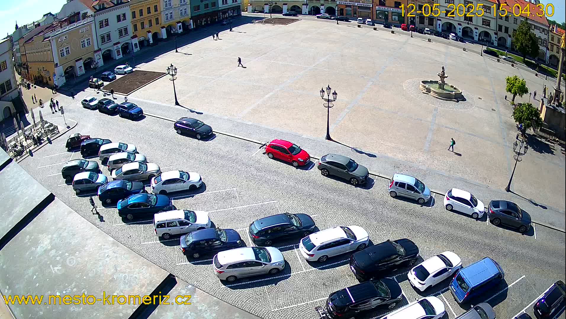 A large, paved town square, under sunny conditions, is partially surrounded by buildings and contains several parked cars and a fountain in the center.