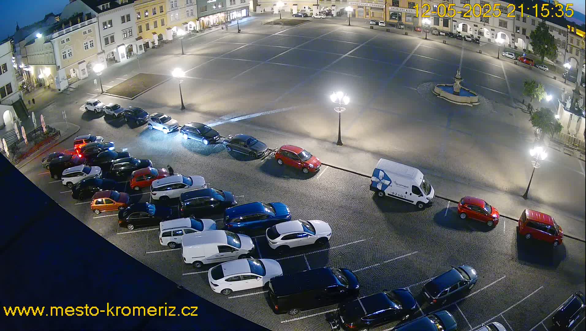 A nighttime, high-angle view of a town square with numerous cars parked along the perimeter, under streetlights.