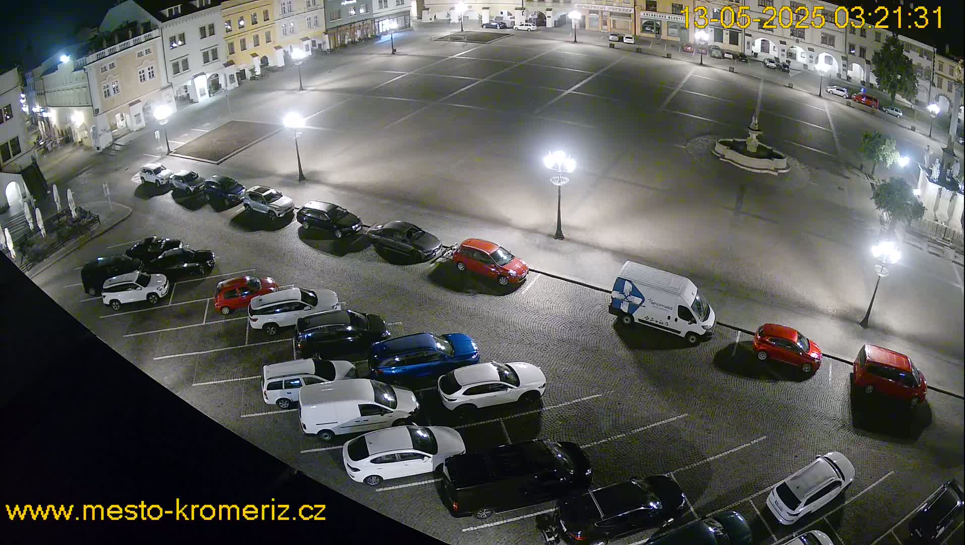 A nighttime, overhead view of a town square with cars parked along the edges and a fountain in the center.