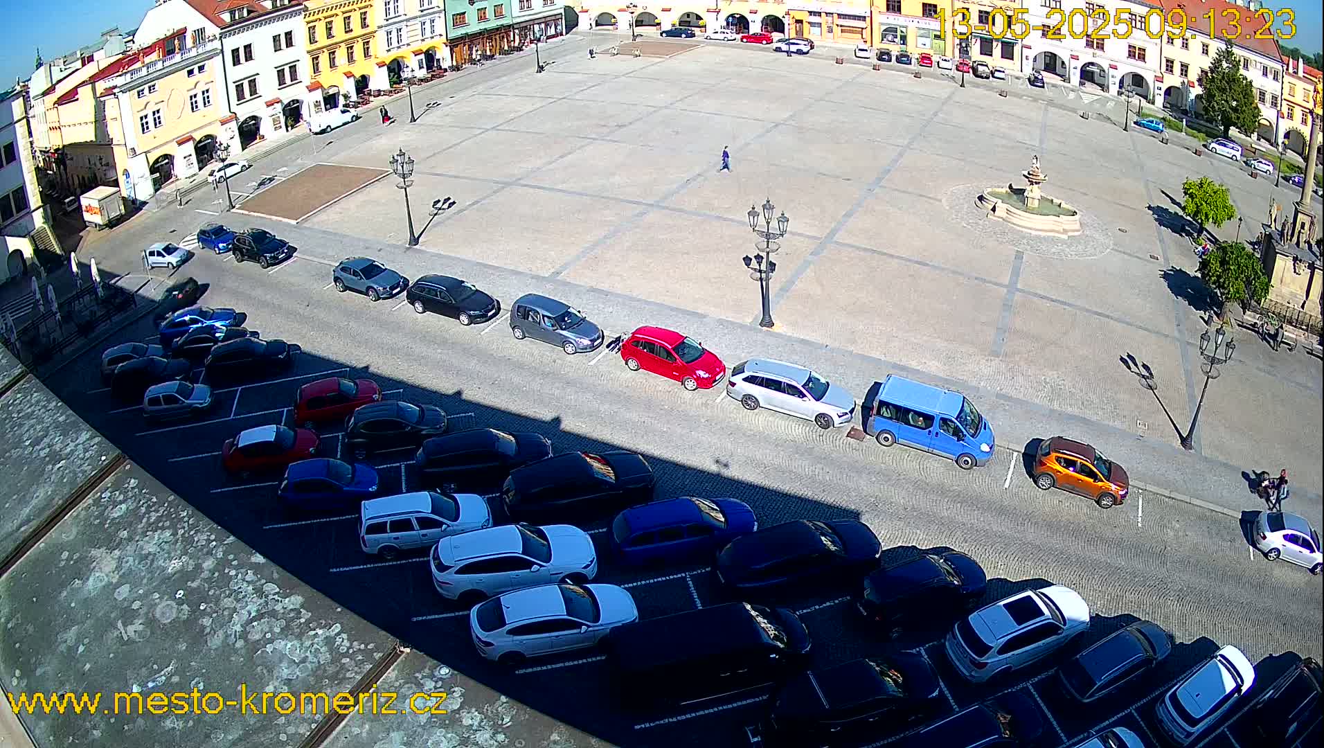 A sunny day reveals a large town square with cars parked along its sides, a fountain at its center, and a few pedestrians strolling about.