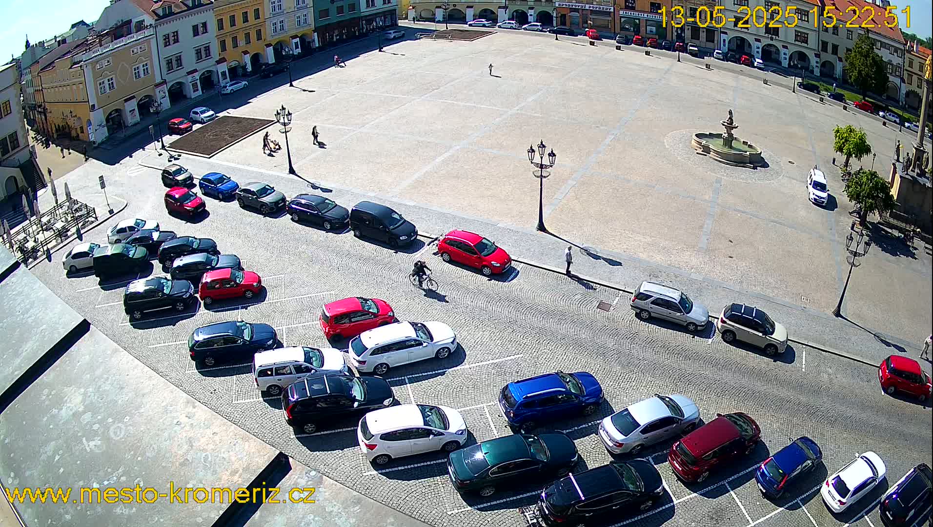 A paved town square with a fountain in the center is surrounded by buildings and numerous cars parked along the edges, under sunny conditions.