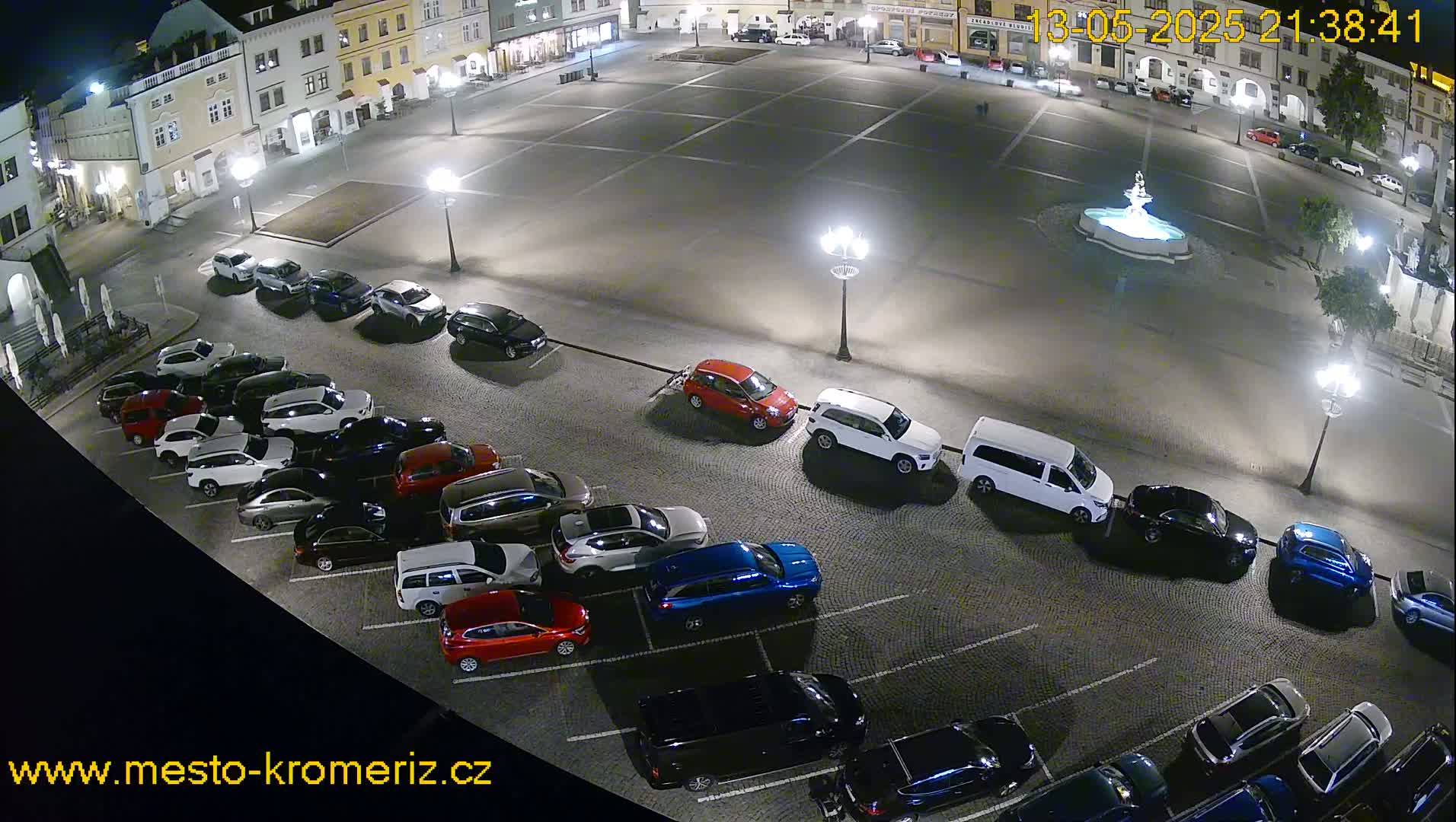 A nighttime aerial view shows a town square with a fountain and several cars parked along the sides of the square.