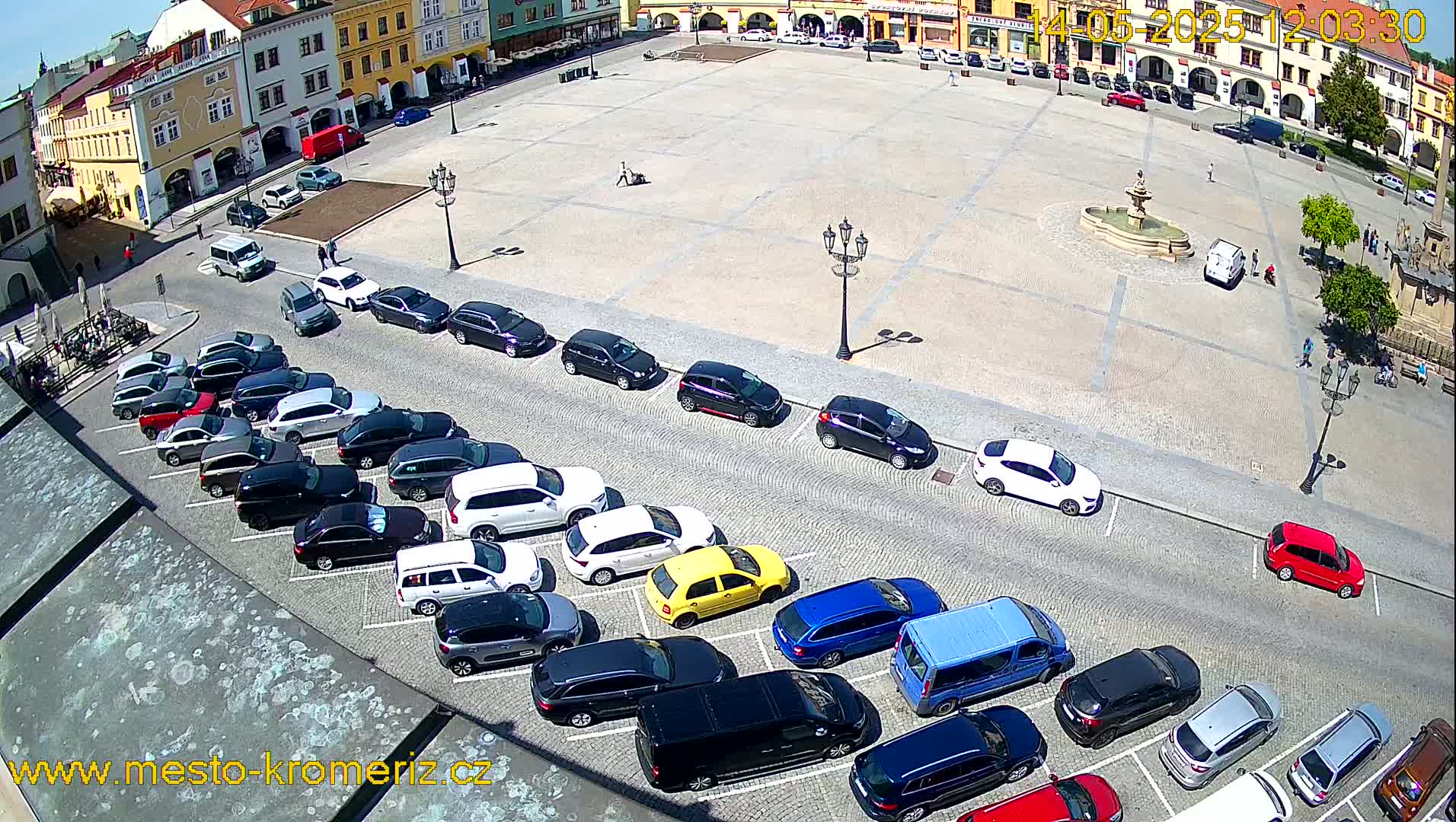 A large town square on a sunny day with numerous cars parked along its edges, a fountain in the center, and buildings surrounding the perimeter.