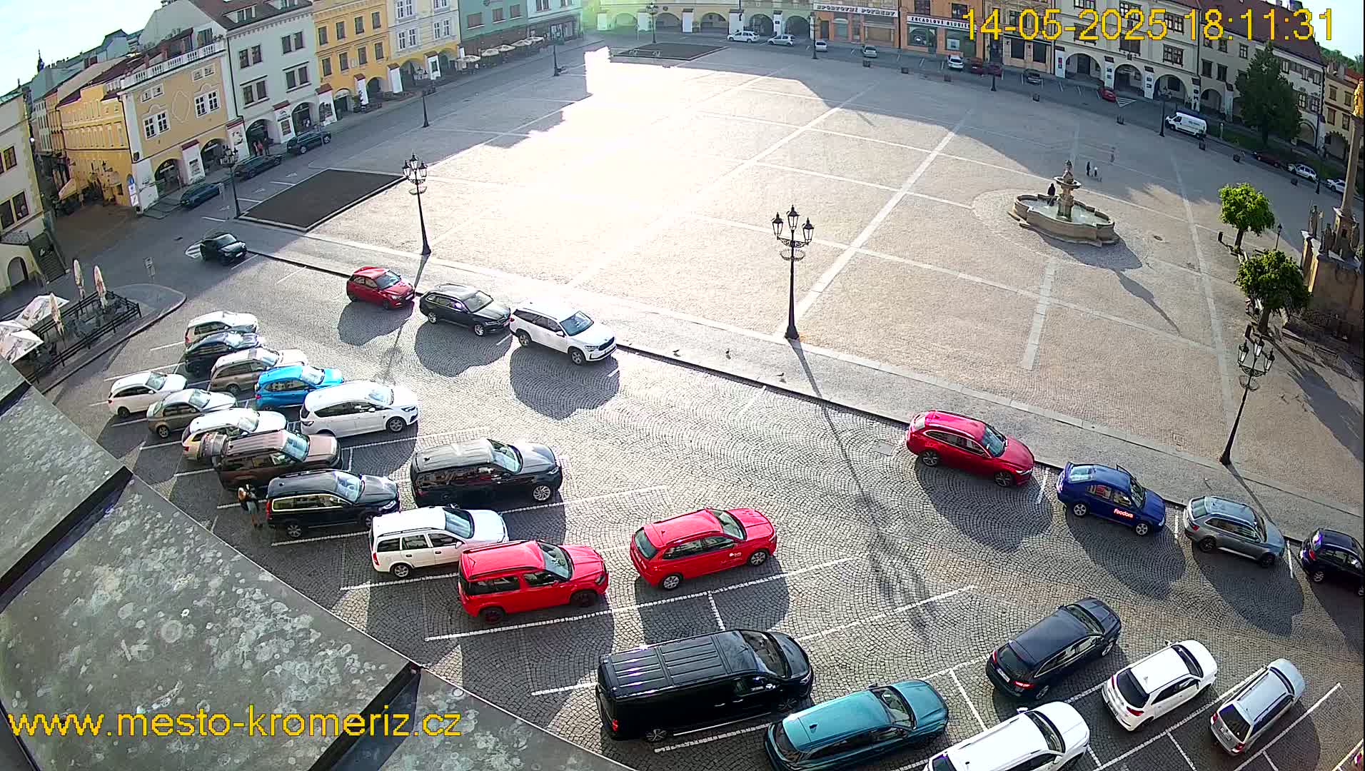 A sunny day shows a large town square with a fountain in the center and numerous cars parked along the edges.