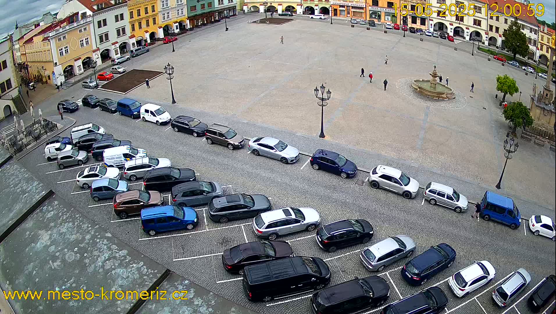 A town square with a fountain in the center, surrounded by parked cars and buildings, under an overcast sky.