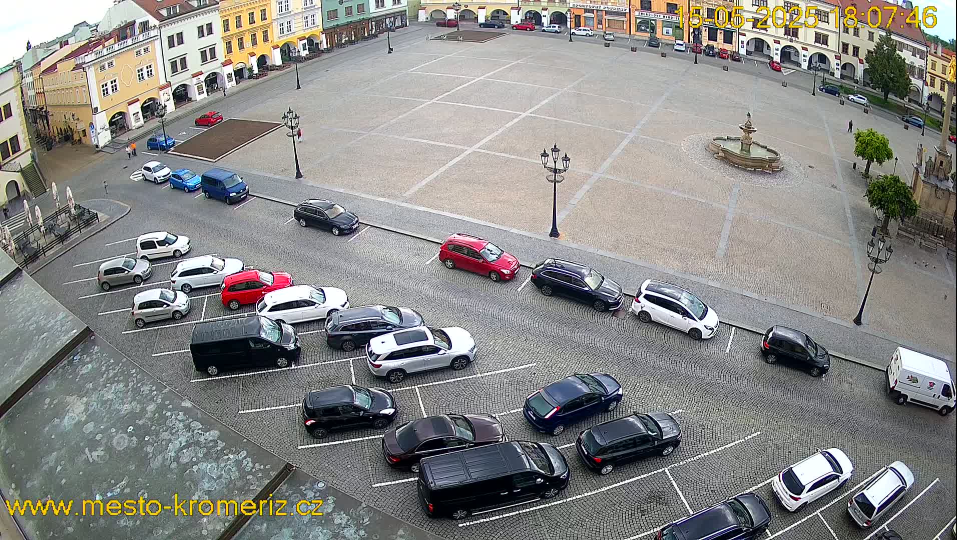 A town square with a fountain in the center, surrounded by parked cars and buildings, on an overcast day.