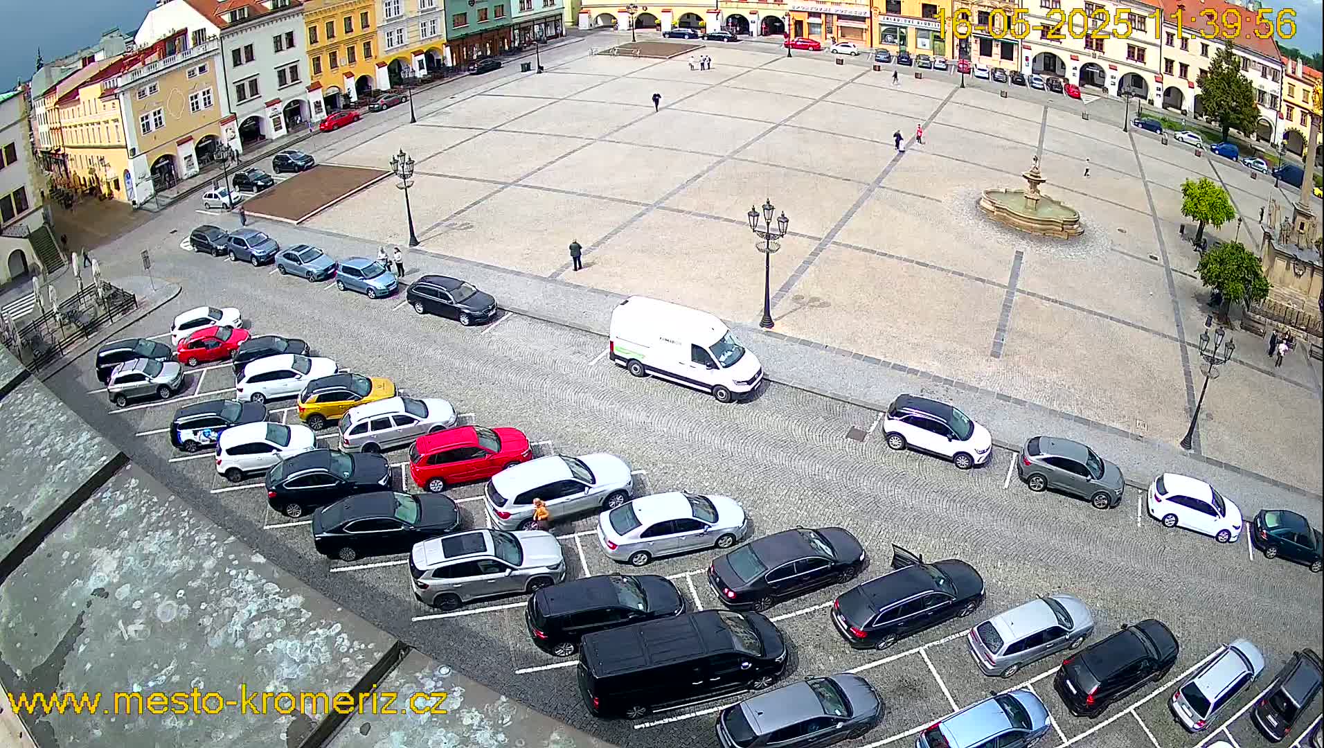An aerial view of a town square with a fountain, surrounded by parked cars and buildings on a partly cloudy day.