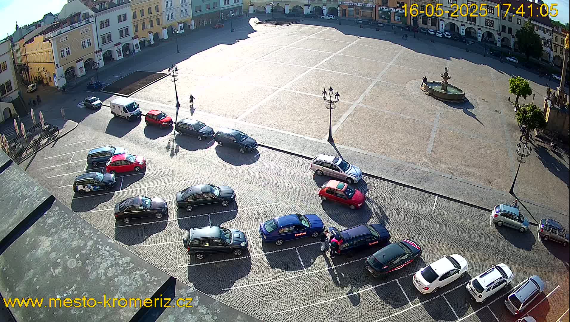 A sunny day reveals a town square with several cars parked along its edge, a fountain in the center, and buildings surrounding the perimeter.