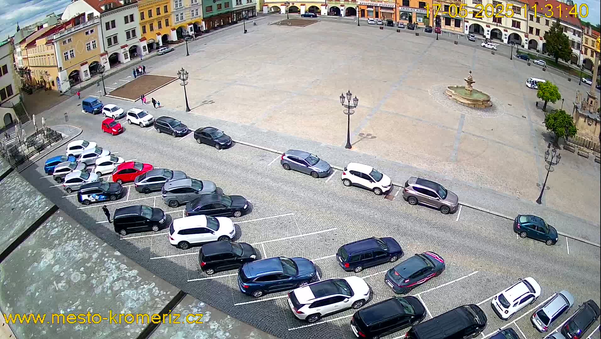 A large town square with a fountain in the center is surrounded by buildings and numerous cars parked along the sides under a partly cloudy sky.