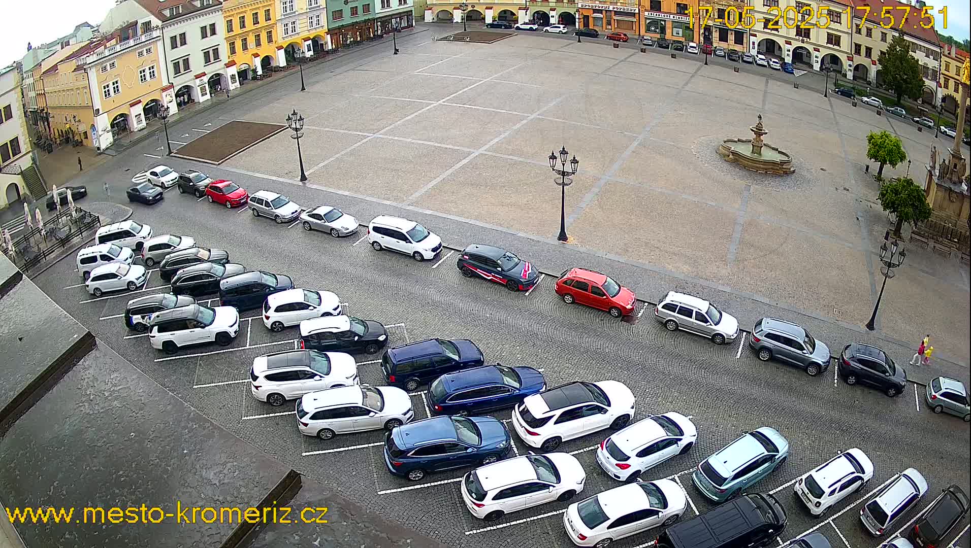 A large town square with a fountain at its center is surrounded by buildings and numerous cars parked along its edges on an overcast day.