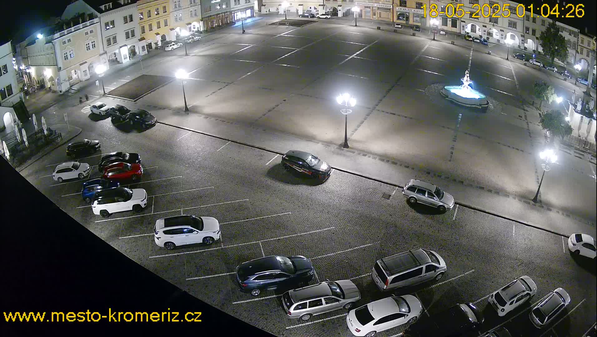A nighttime aerial view of a town square's parking lot and surrounding buildings, featuring several cars parked and a fountain in the center of the square.