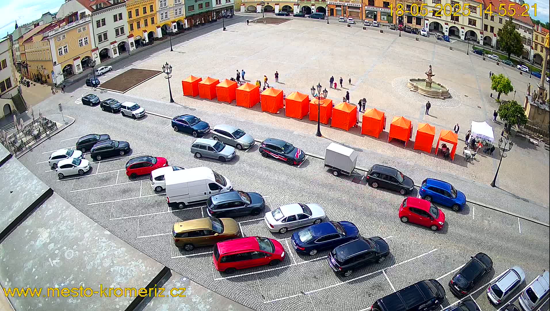 A high-angle view shows a town square on a sunny day with a row of orange tents, a fountain, and numerous cars parked in a lot adjacent to the square.