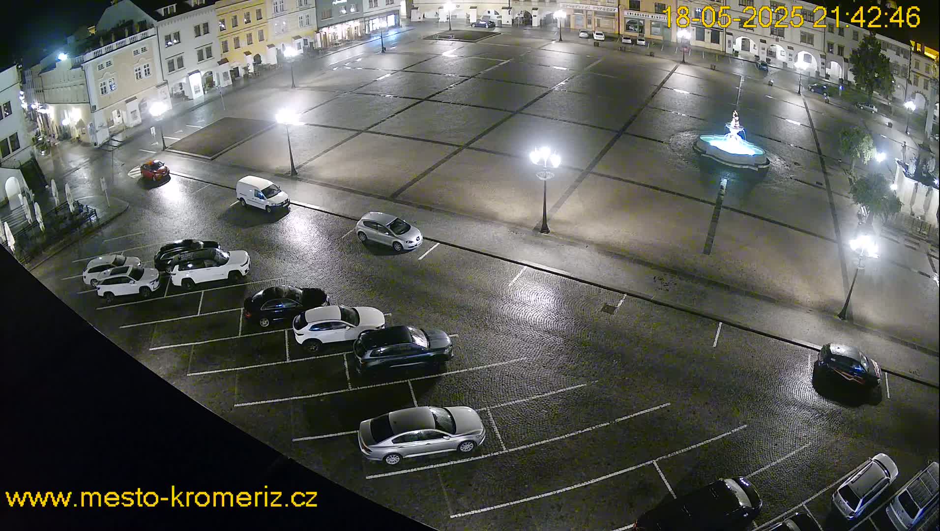 A nighttime view of a town square with a fountain and several cars parked in a lot, under a wet, dimly lit sky.