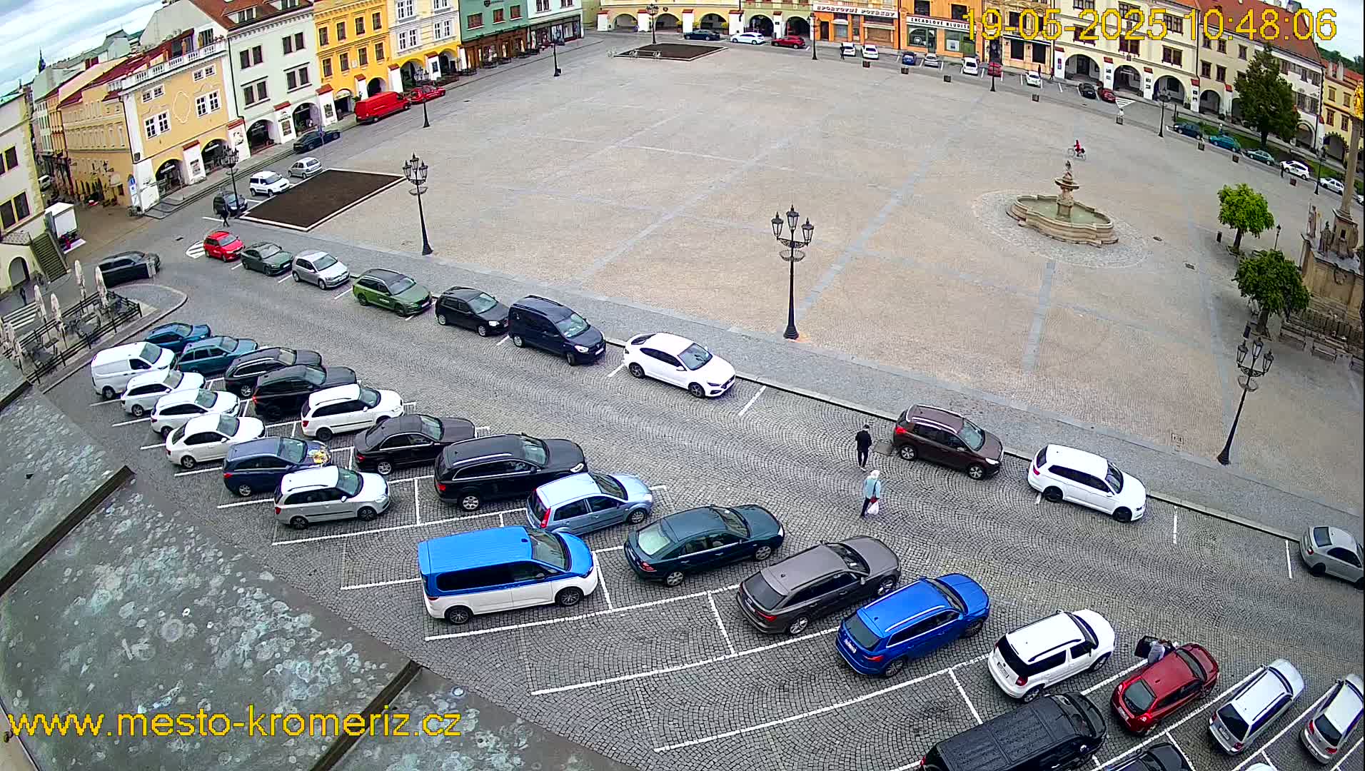 An overhead view shows a town square with a fountain in the center, surrounded by buildings and numerous cars parked in adjacent lots, under a cloudy sky.