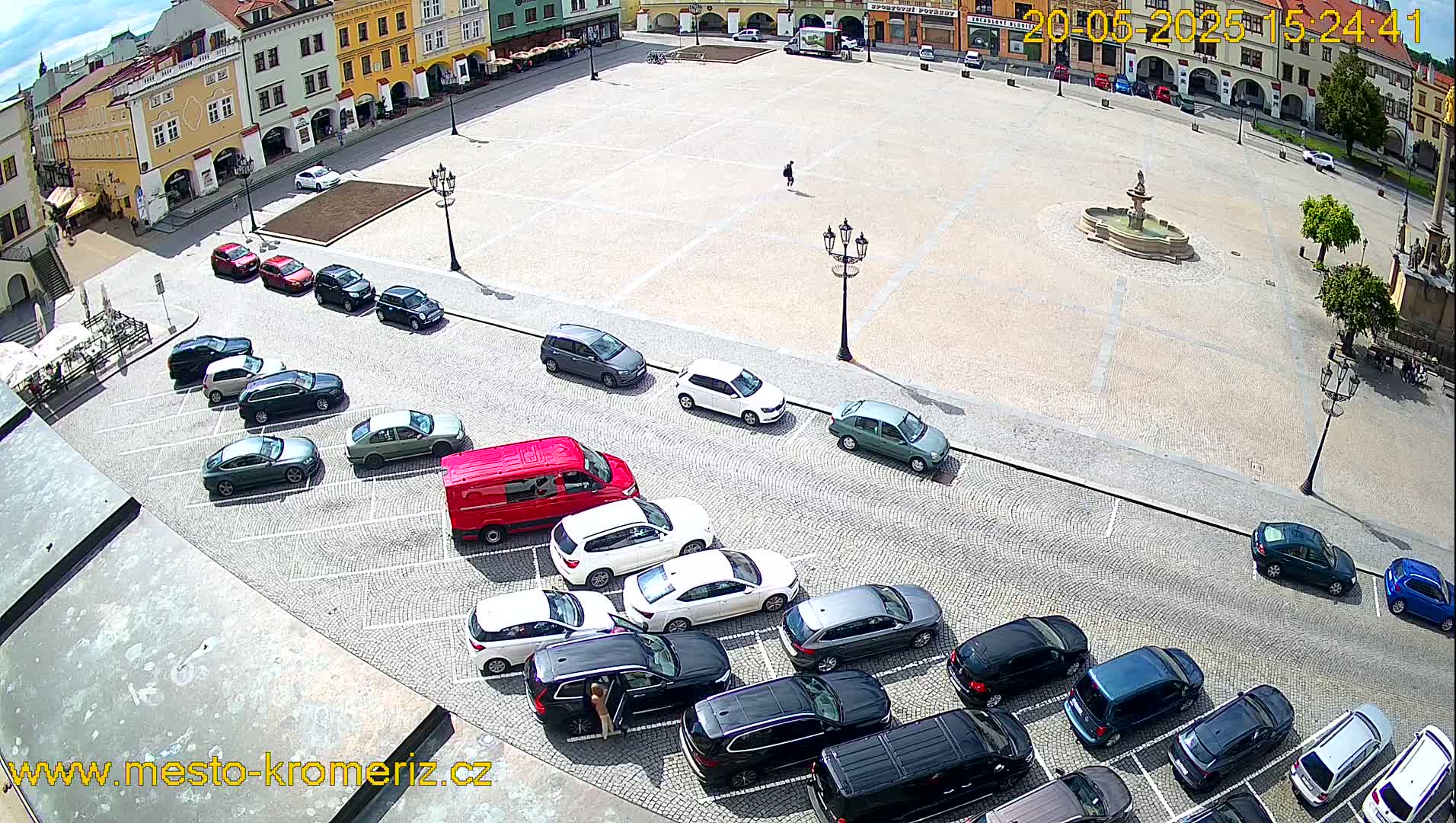 A large town square with a fountain in the center is surrounded by parked cars and buildings on a sunny day.