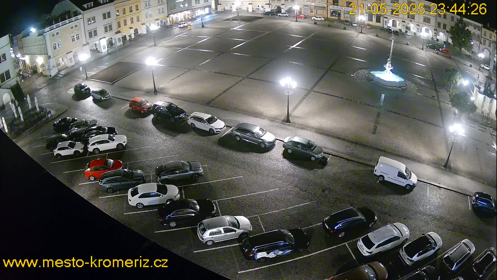 A nighttime, wet town square with a fountain in the center and several cars parked along the edges.