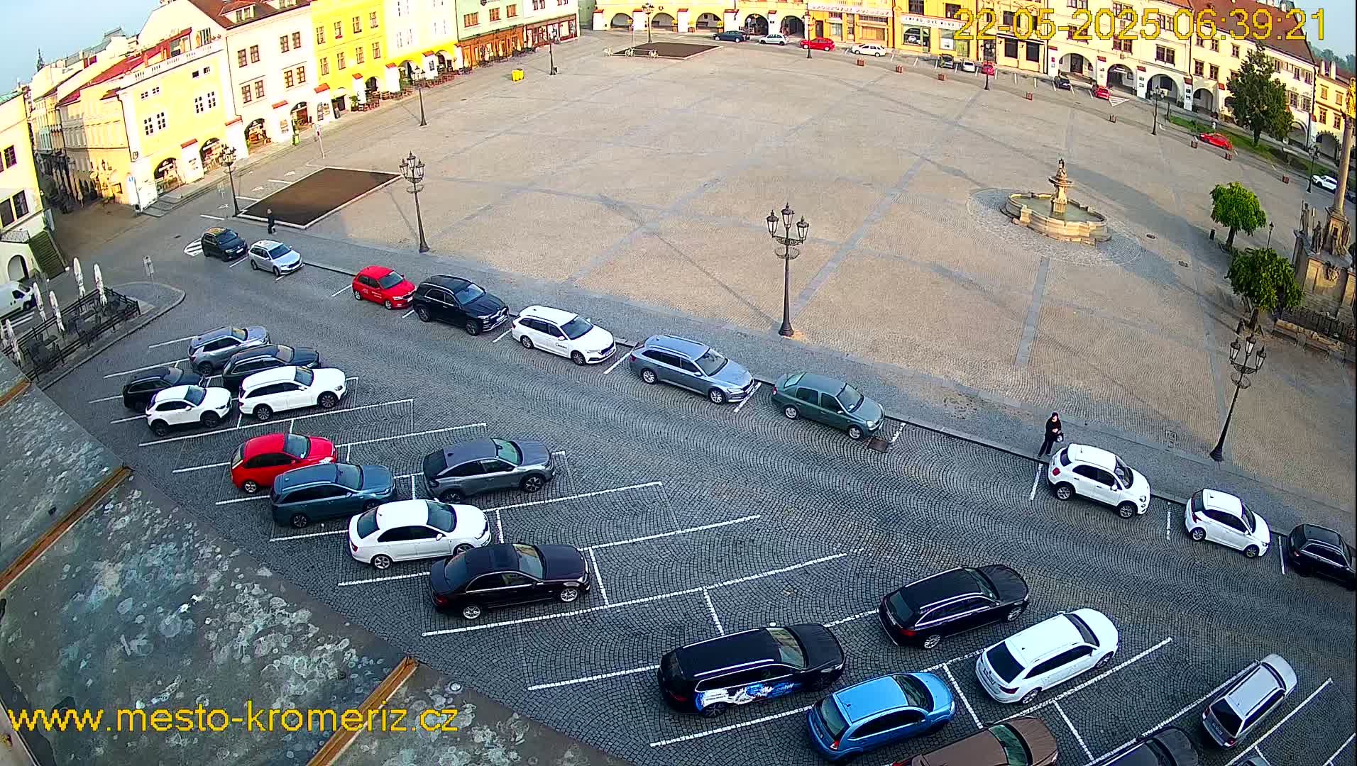 A large, paved town square with a fountain in the center, surrounded by buildings, and several cars parked in a lot adjacent to the square on a sunny day.
