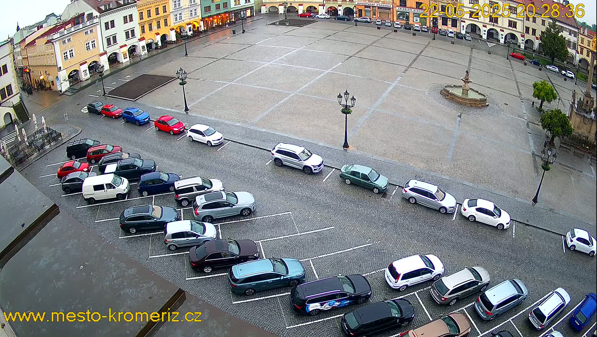 A large, cobblestone town square with a fountain in the center is surrounded by buildings and several cars parked along its edge on a wet, overcast day.