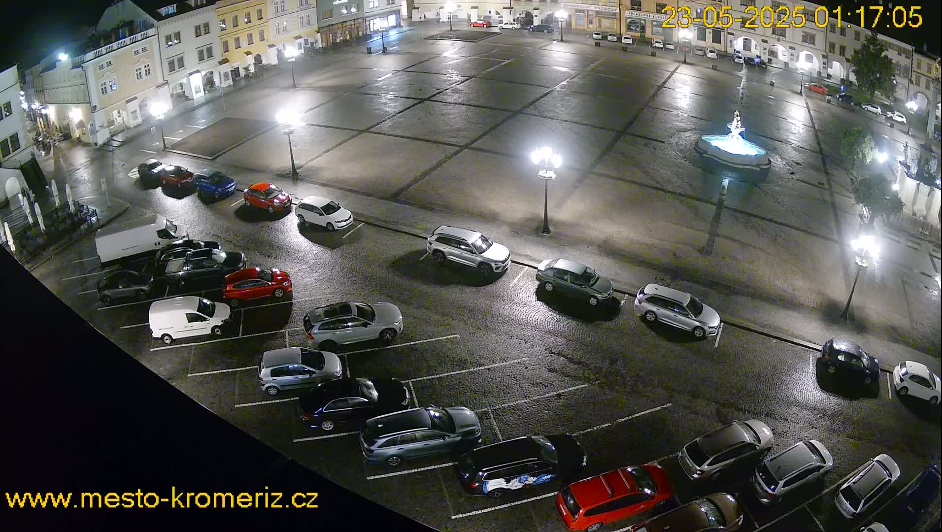 A nighttime, wet town square shows a fountain and numerous cars parked in the surrounding lots, illuminated by streetlights.