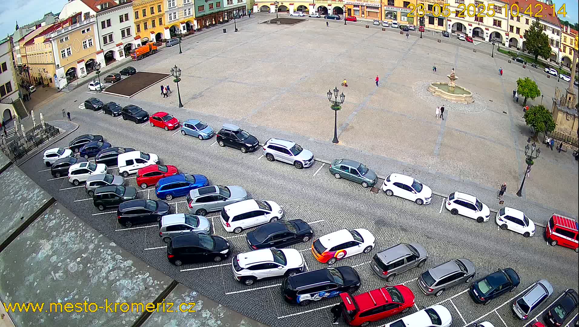 A mostly empty town square with a fountain in the center, surrounded by parked cars and buildings under a partly cloudy sky.