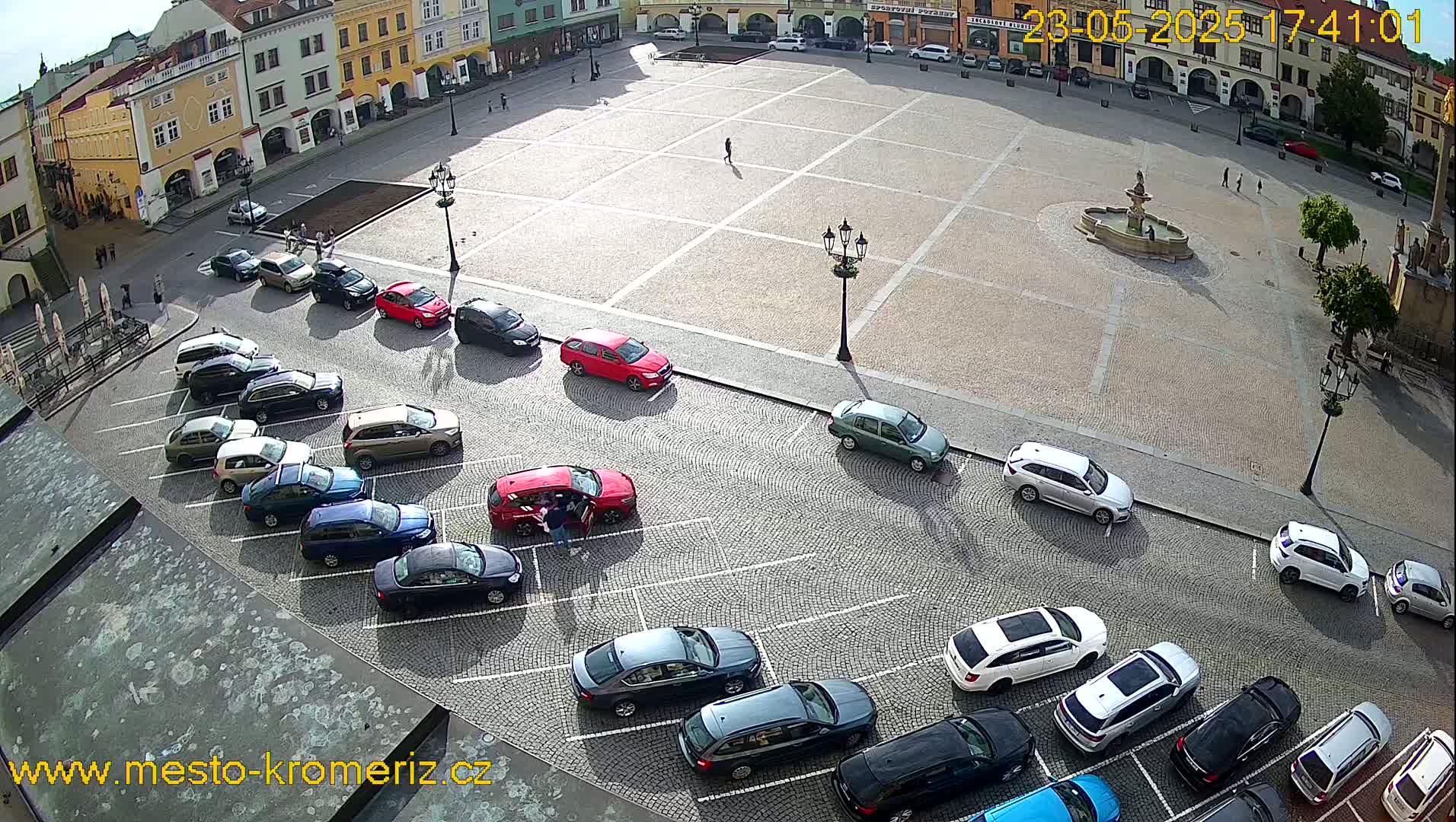 An aerial view of a town square with parked cars lining the perimeter, a fountain in the center, and several pedestrians scattered around under a sunny sky.