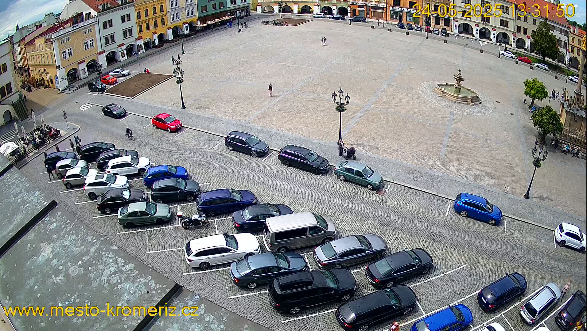 A large town square with a fountain in the center is surrounded by buildings, and several cars are parked along the perimeter on a partly sunny day.