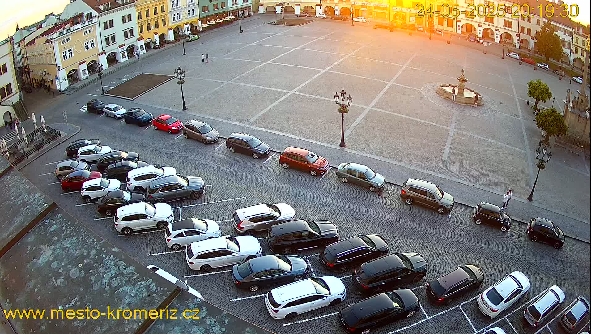 A paved town square at sunset, with cars parked along its edges and a fountain in the center.