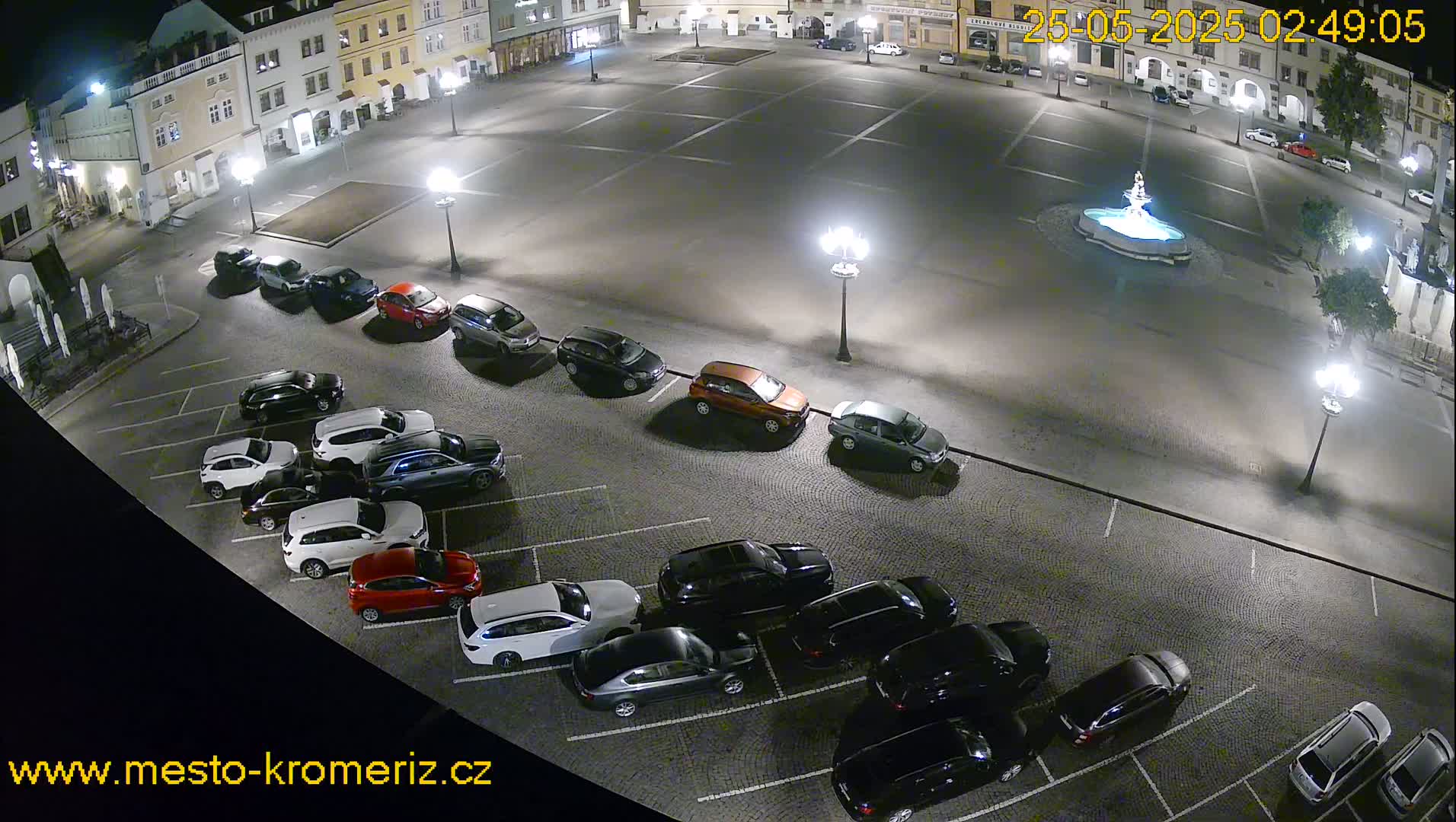 A nighttime aerial view of a town square with a fountain at its center, surrounded by parked cars and illuminated by streetlights.