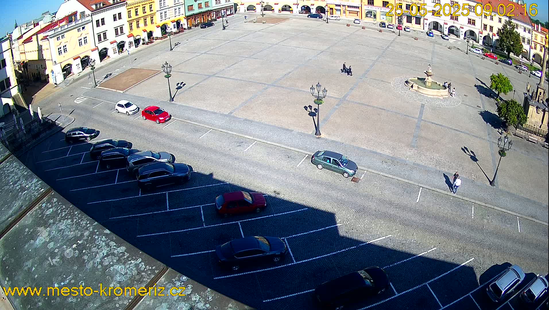 A sunny day reveals a large town square with a fountain at its center, surrounded by colorful buildings and bordered by a parking lot filled with cars.