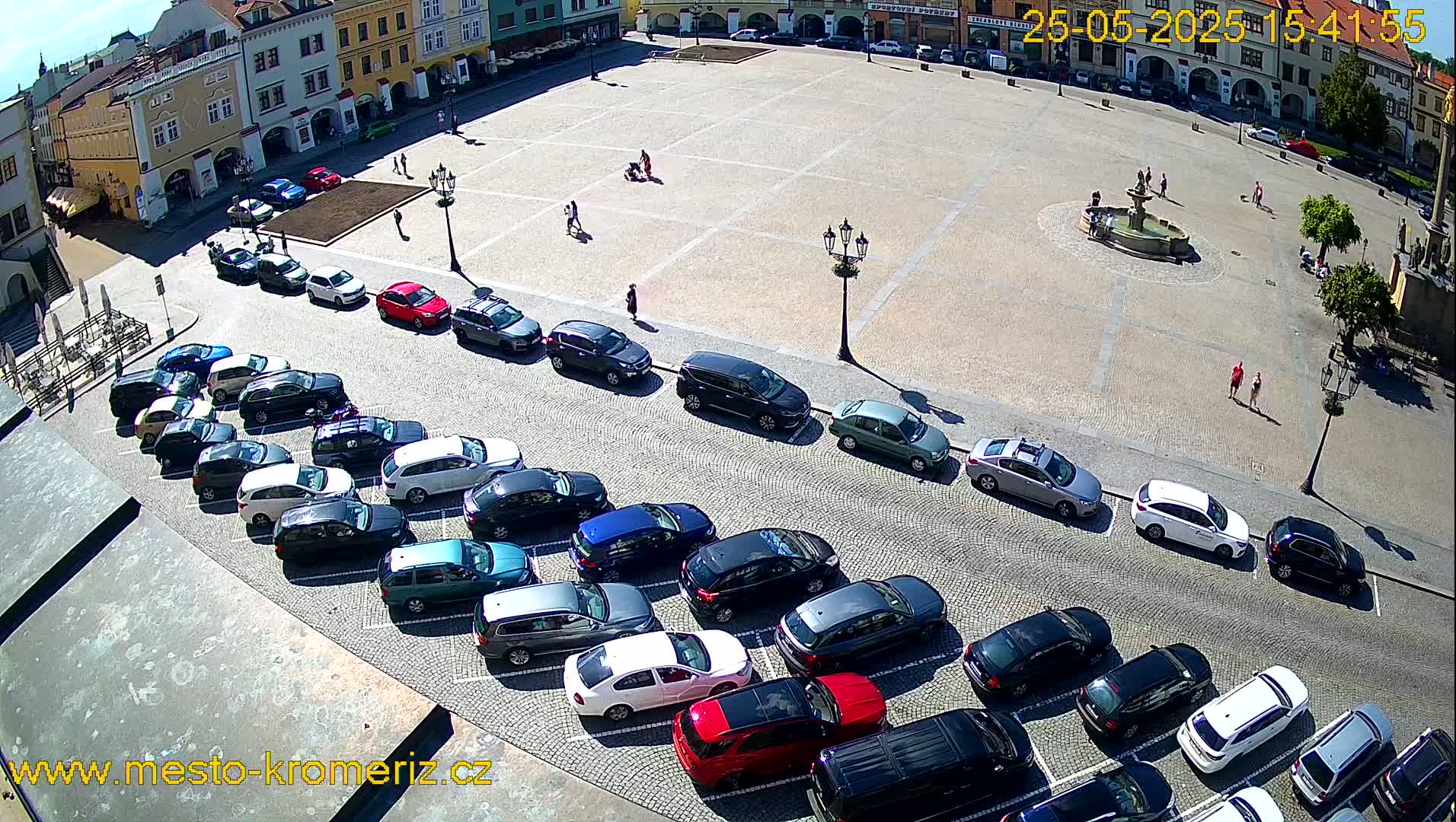 A sunny day in a town square shows a fountain, several pedestrians, and numerous cars parked along the perimeter.