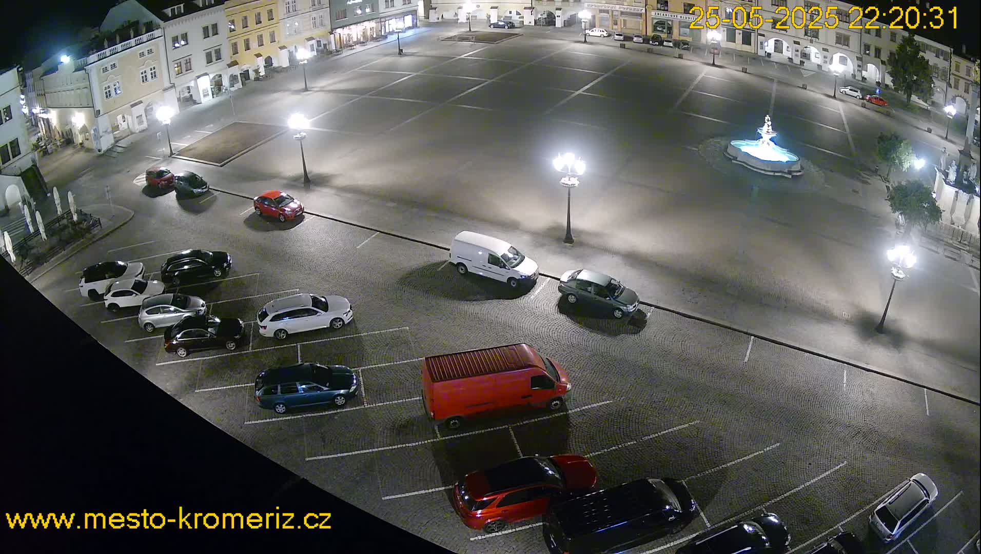A nighttime aerial view of a town square with several cars parked in a lot and a fountain in the center, illuminated by streetlights.