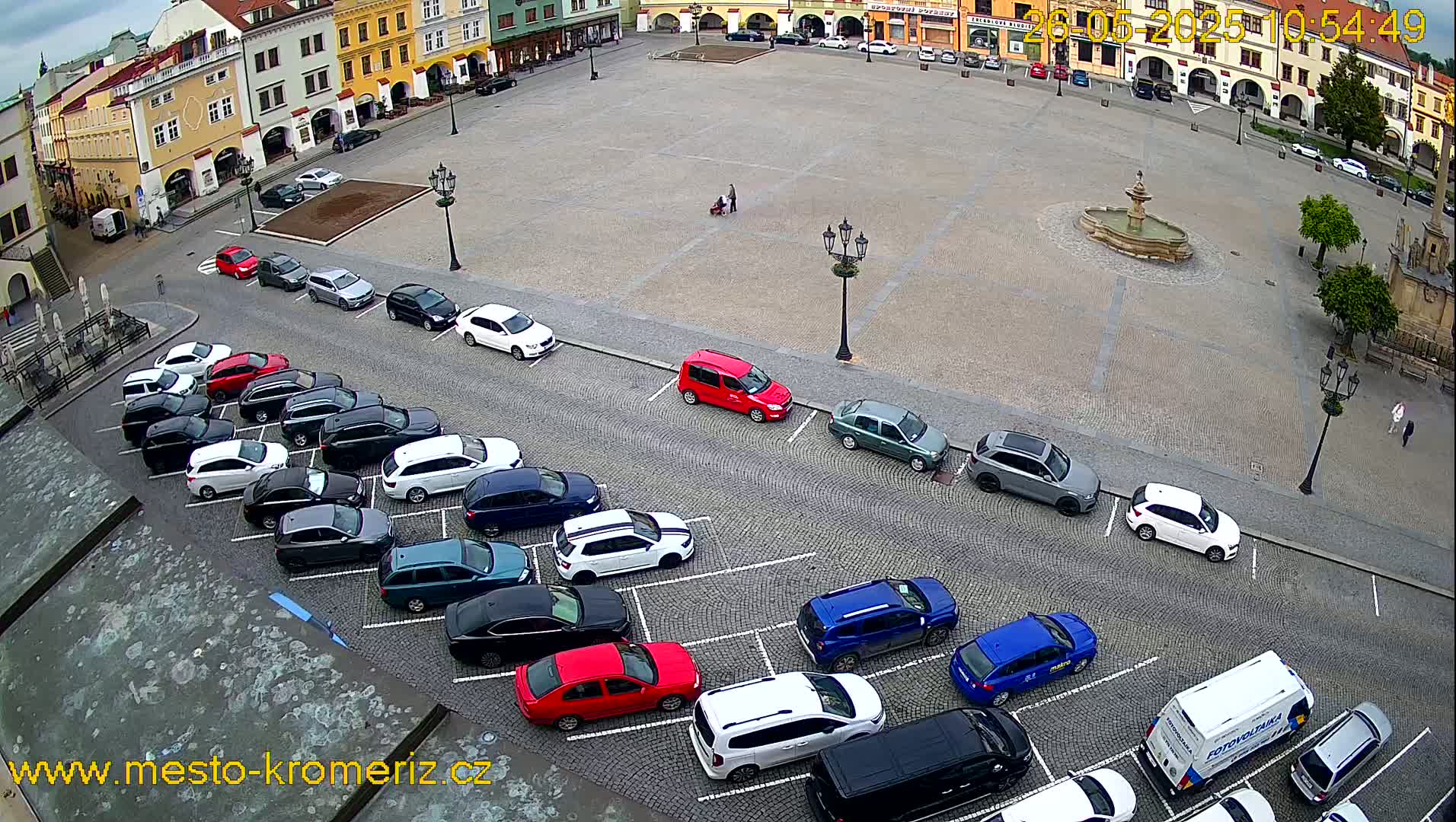 An overcast day reveals a town square with a fountain in the center, surrounded by buildings and cars parked in two adjacent lots.