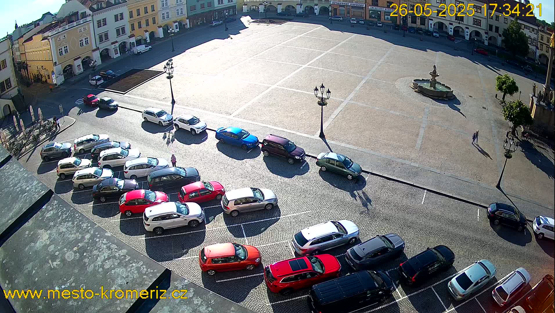 A sunny day shows a large town square with a fountain and several cars parked along the edge.