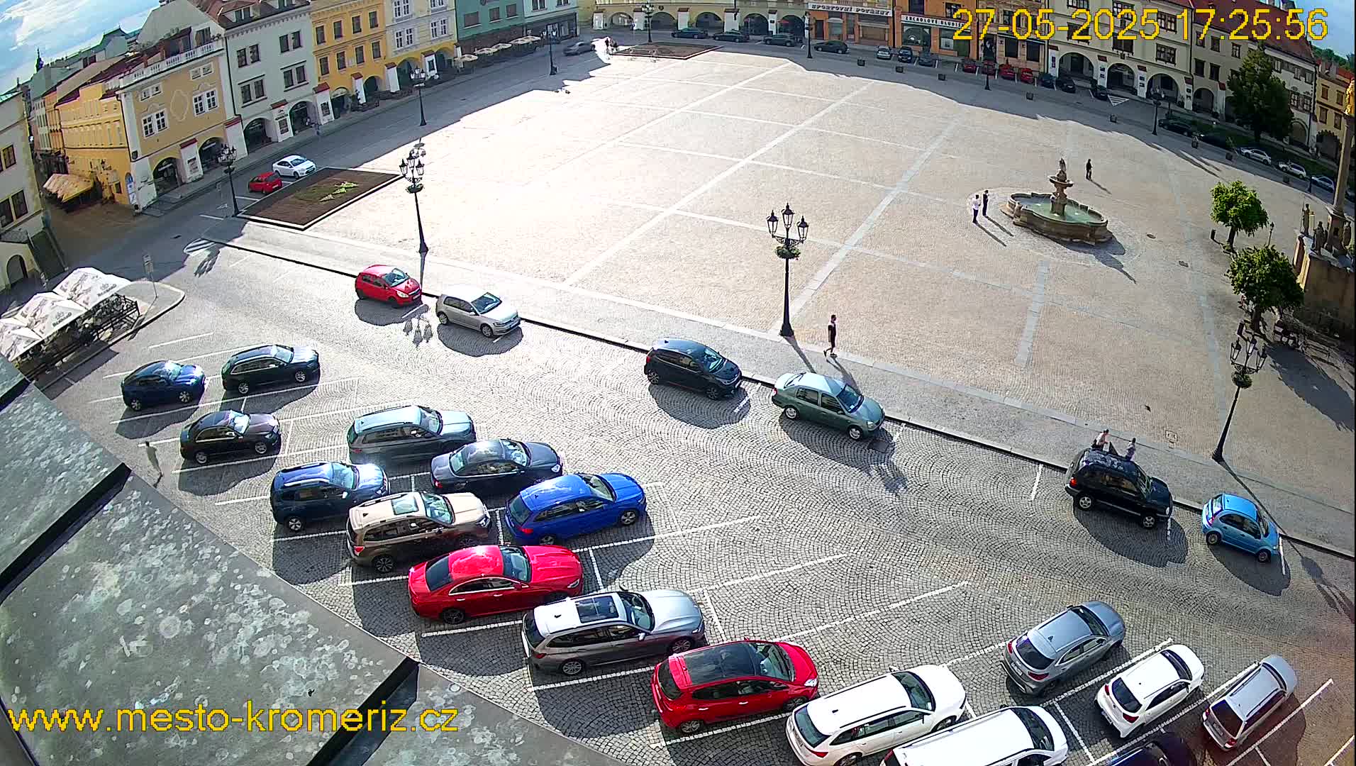 A cobblestone town square, under sunny conditions, is surrounded by buildings and has several cars parked along the edge.