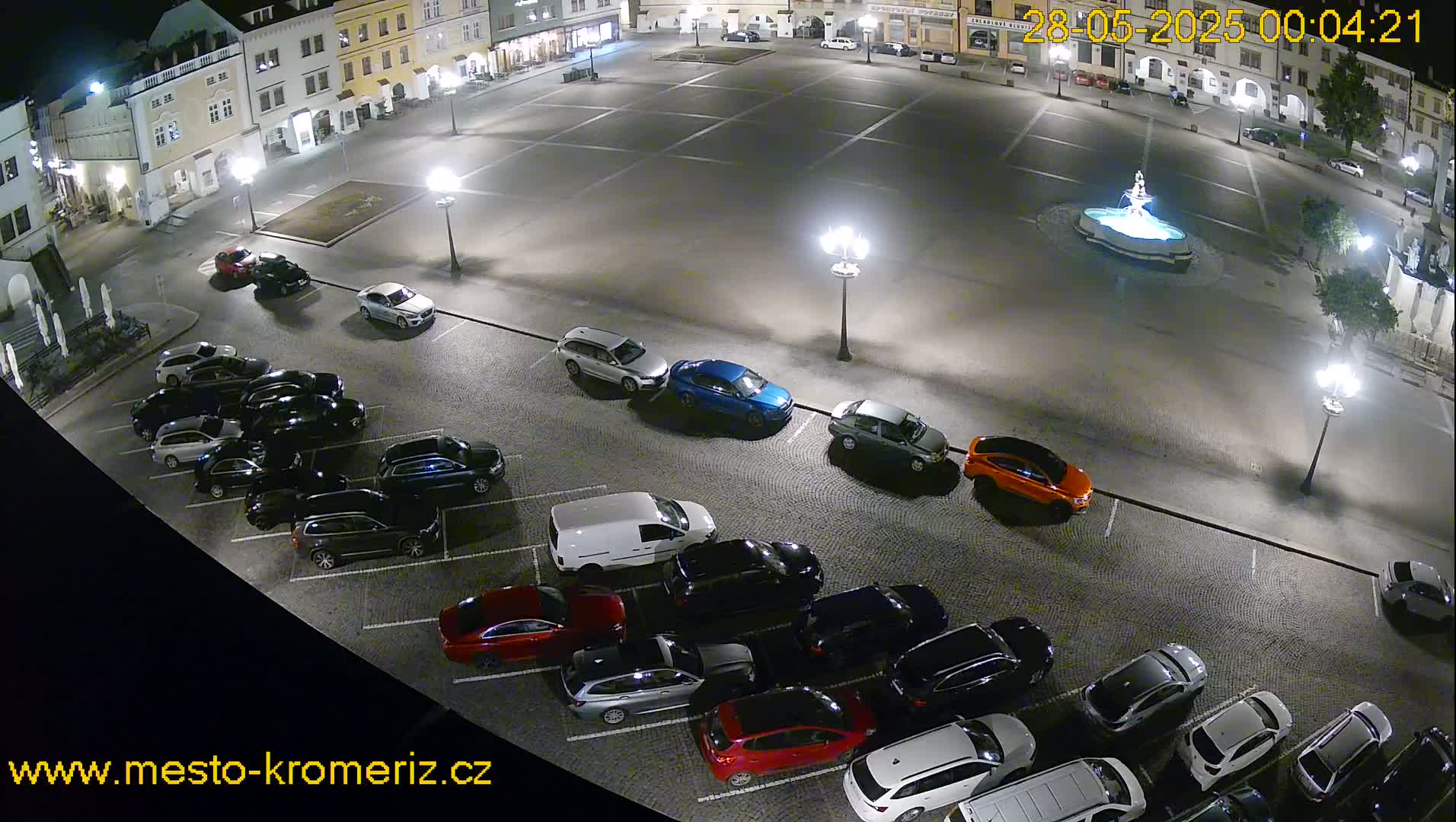 A nighttime, high-angle view of a town square with a fountain and numerous cars parked in a lot adjacent to the square.
